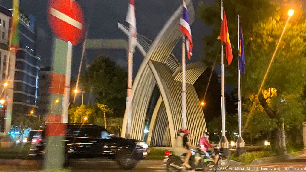 Angular illuminated gateway beside a Ho Chi Minh City street with flags, trees, cars, and passing motorbikes at night.