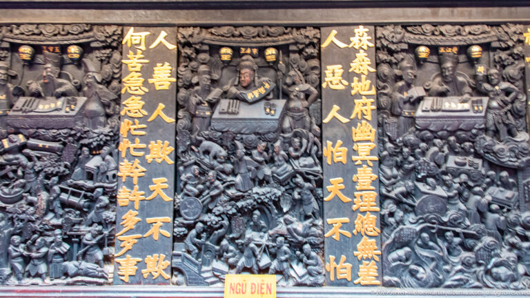 Dark carved temple relief with seated figures, gilded Chinese inscriptions, cloud motifs, and a small red “Ngũ Điện” label.