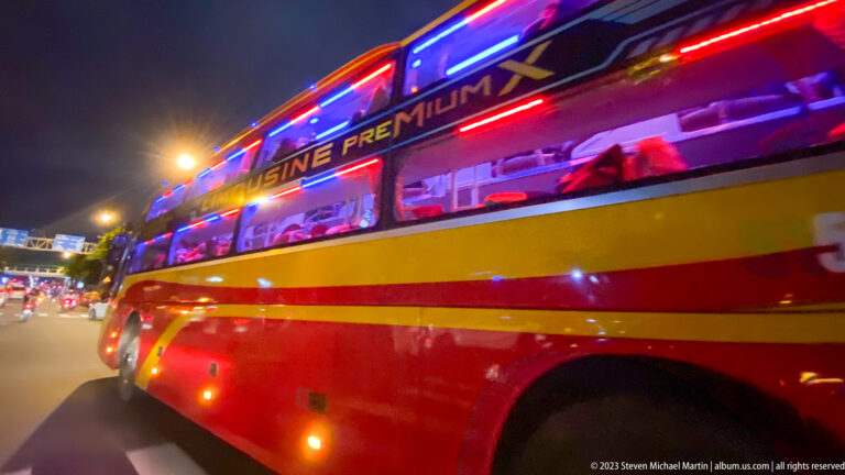 Red-and-yellow premium coach with bright blue and red neon lighting traveling through Ho Chi Minh City at night.