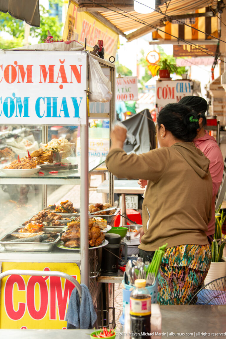 Vietnamese street-food stall with signs for cơm mặn and cơm chay, prepared dishes, a seated vendor, and sidewalk customers.