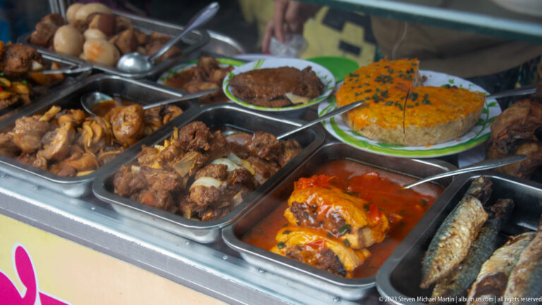 Close view of metal trays holding braised meats, grilled fish, stuffed vegetables, and prepared rice plates at a food stall.