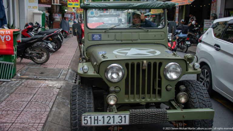 Olive-drab open-top jeep with white star, round headlights, rugged tires, and Vietnamese license plate parked among city vehicles.