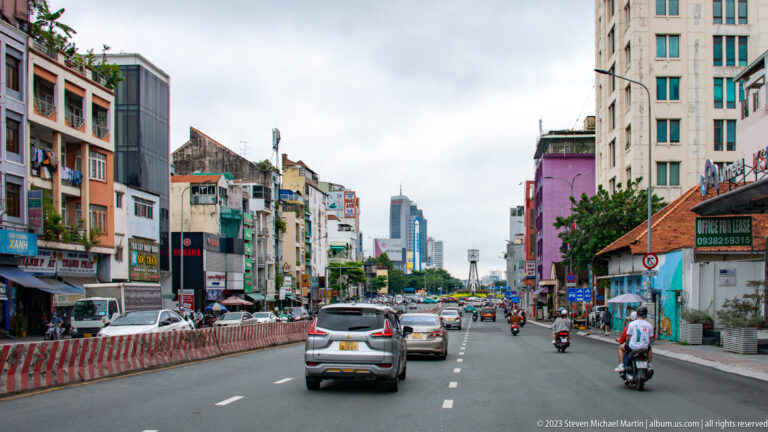 Wide Ho Chi Minh City avenue with cars and motorbikes, low-rise shops, apartment façades, and high-rise towers in the distance.