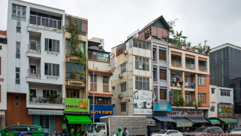 Row of narrow Ho Chi Minh City shophouses with enclosed balconies, shop signs, rooftop plants, and varied façades.
