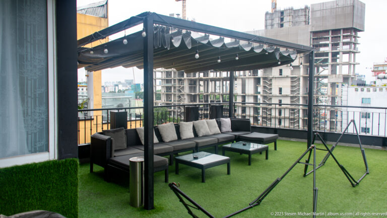Covered rooftop lounge with gray sectional seating, string lights, swing frames, artificial turf, and city buildings beyond.
