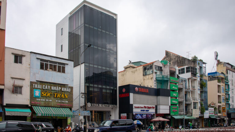 Dark-glass mid-rise office building flanked by older narrow shophouses with signs, windows, and weathered façades.