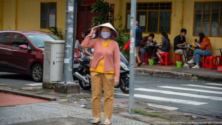 Woman in a conical hat and face mask crossing the street beside diners seated on low red stools.