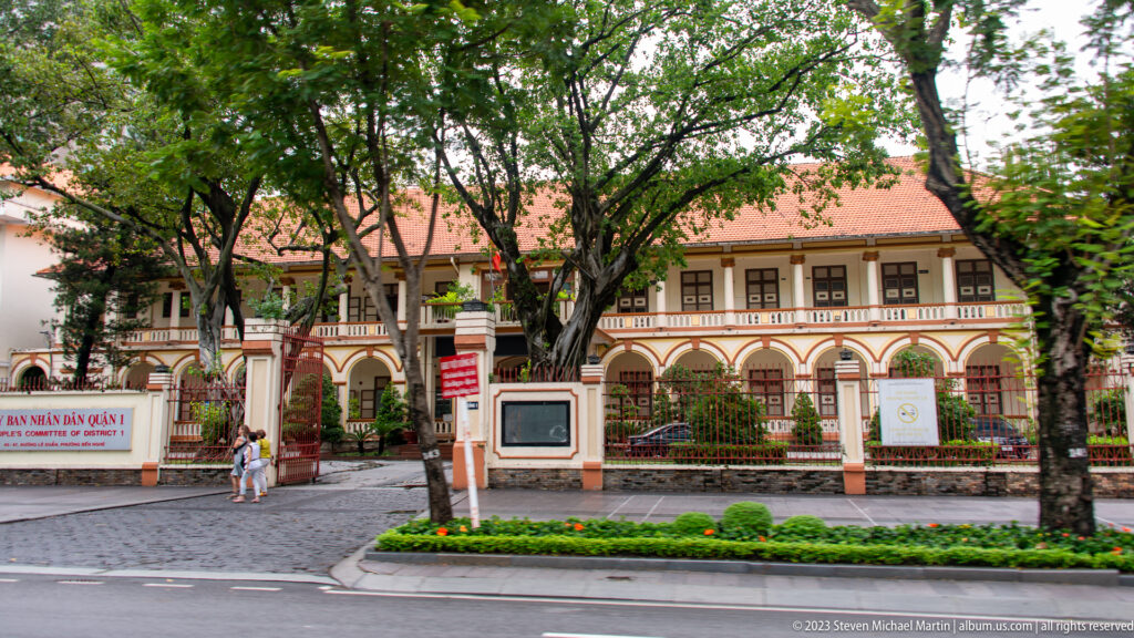 Cream institutional building with red tile roof, arcaded upper level, trees, gated frontage, and a landscaped street garden.
