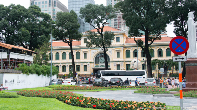 Yellow colonial-style public building with arched windows and red roof beyond flower beds, trees, tour vehicles, and pedestrians.