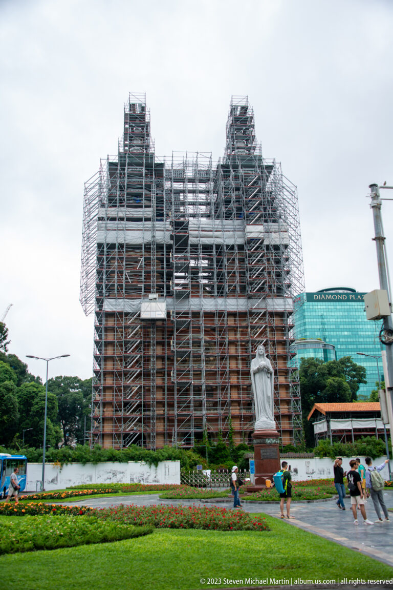 Notre-Dame Cathedral Basilica of Saigon covered in dense scaffolding, with its twin towers visible above a landscaped square.