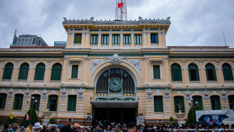 Yellow façade of Saigon Central Post Office with central clock, arched entrance, green shutters, and crowds below.