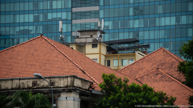 Red tile roofs and weathered low-rise walls in front of a large blue-glass high-rise building.