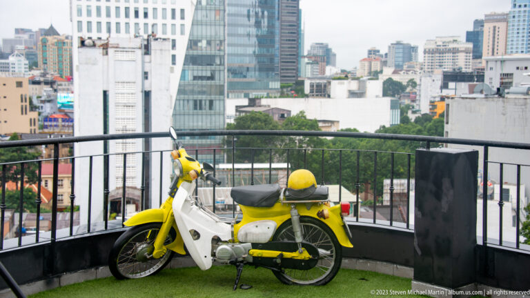 Yellow-and-white vintage step-through motorbike displayed on artificial turf beside a rooftop railing.