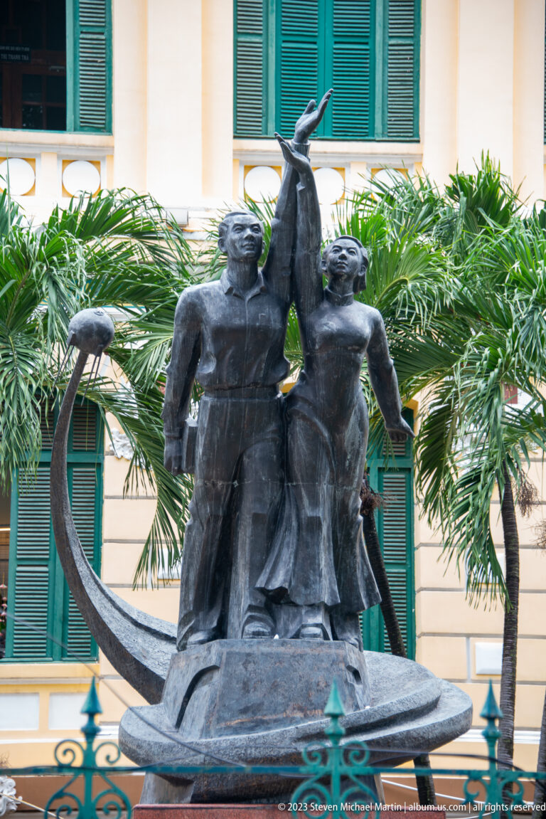 Bronze sculpture of three stylized figures reaching upward, framed by palm trees and a pale building with green shutters.