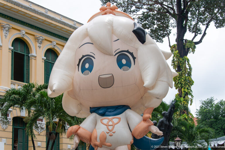 Large white anime-style inflatable mascot with horns and blue neck detail standing outside a historic building.