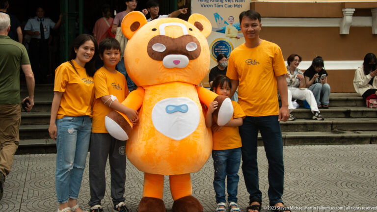 Family in matching yellow shirts posing with a large orange bear-like mascot on outdoor steps.