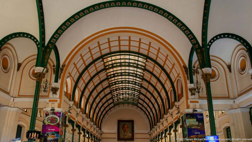 Long vaulted hall of Saigon Central Post Office with green iron ribs, side counters, and a Ho Chi Minh portrait at the far end.
