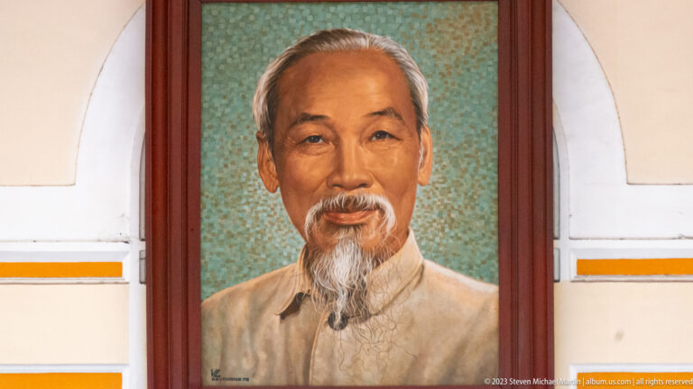 Formal bust portrait of Ho Chi Minh with white beard and pale tunic displayed in an arched interior panel.