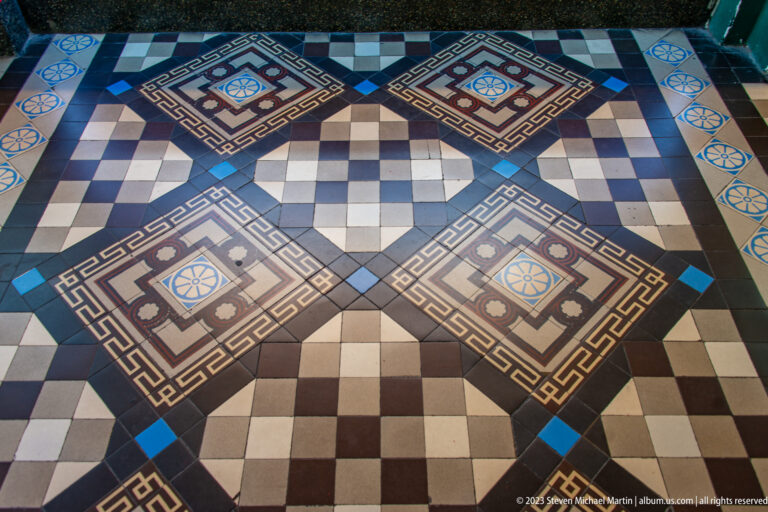 Geometric tile floor in blue, brown, cream, and white with checkerboards, rosettes, diamonds, and Greek-key borders.
