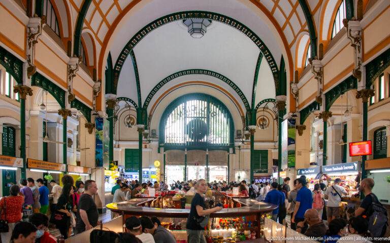 Crowded main hall of Saigon Central Post Office looking toward the large arched entrance under white, green, and orange vaults.