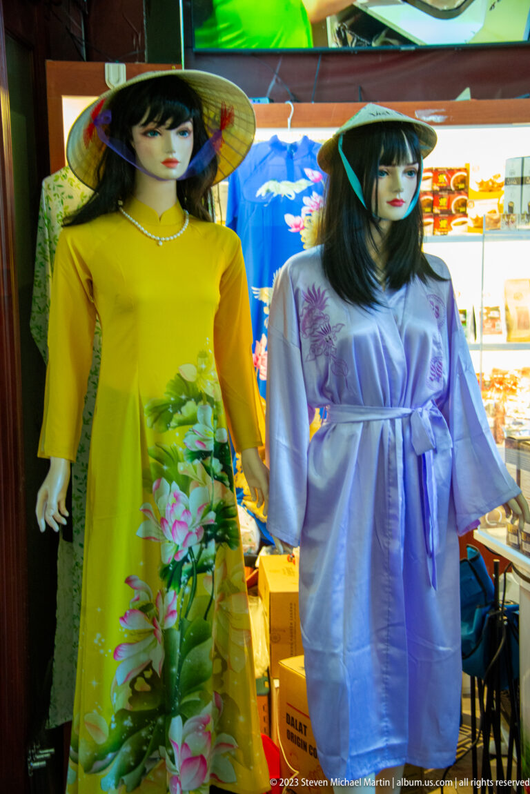 Two mannequins wearing yellow and lavender Vietnamese áo dài with conical hats in a brightly lit shop display.