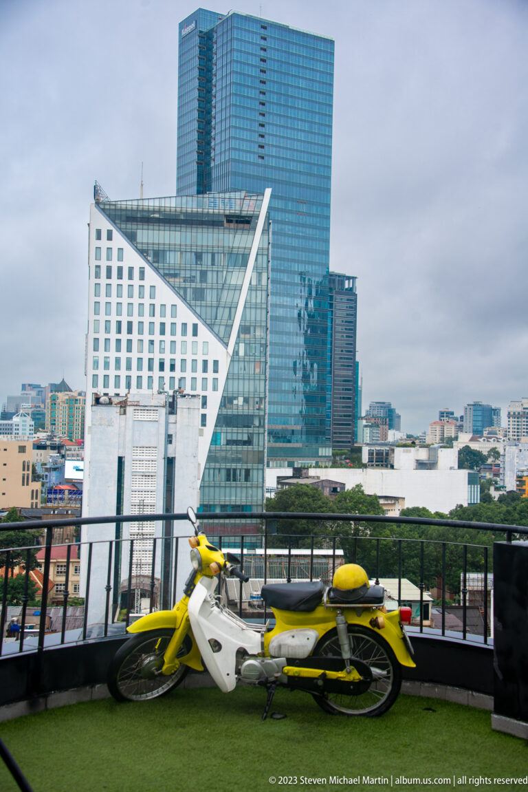 Yellow motorbike on a rooftop terrace below angular white and blue-glass office towers.
