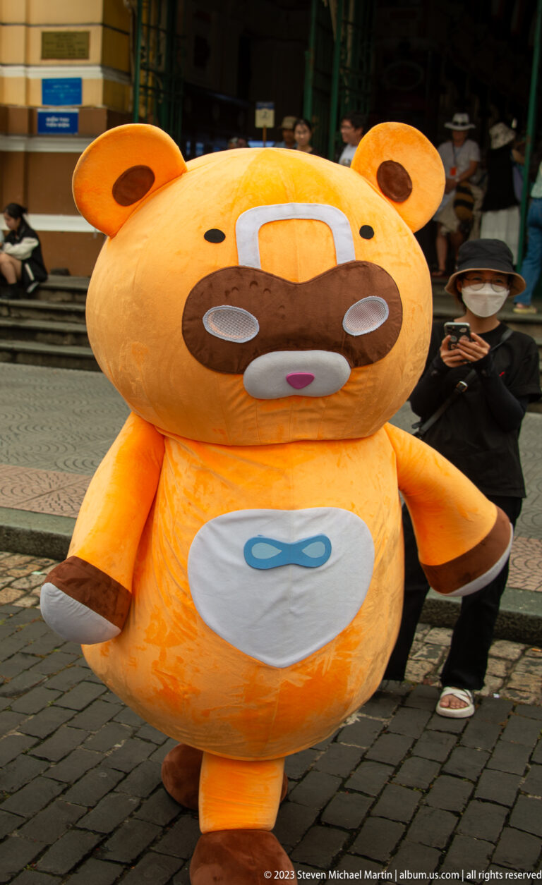 Large orange bear-like mascot with rounded ears and white belly emblem walking past people at an outdoor event.