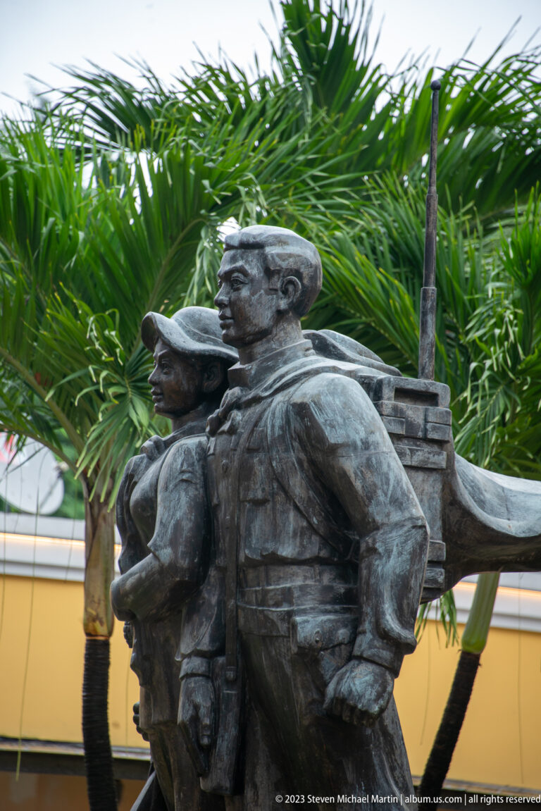 Bronze statue of an armed man beside a woman and child, framed by palm foliage.