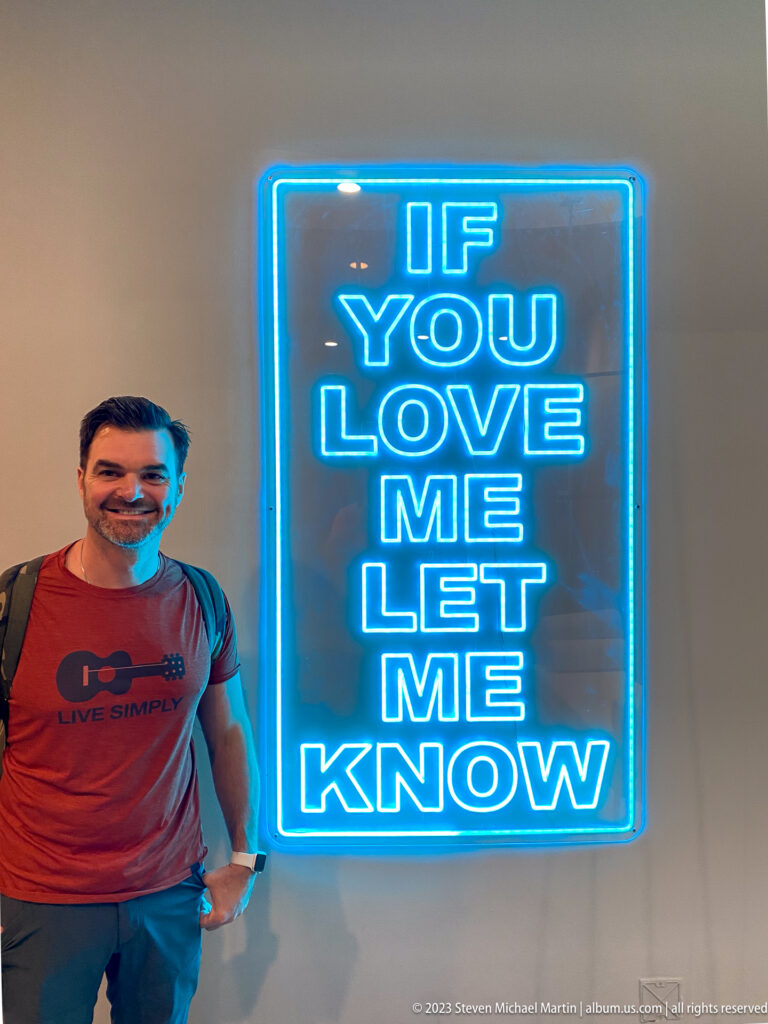 Smiling man beside blue neon words reading, “If you love me let me know.”