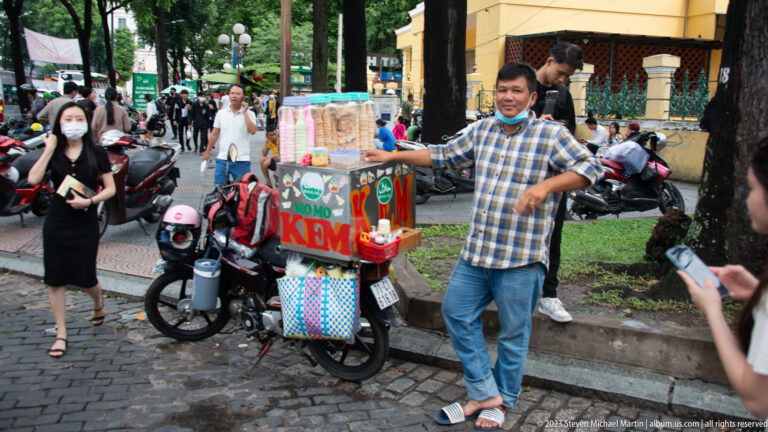 Vendor beside a colorful “Kem” ice-cream cart among motorbikes and pedestrians.
