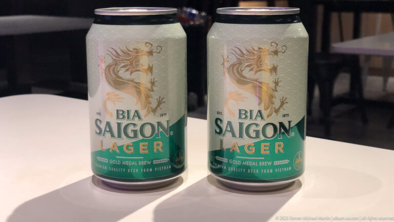 Two cream-and-green Bia Saigon Lager cans standing together on a white table.