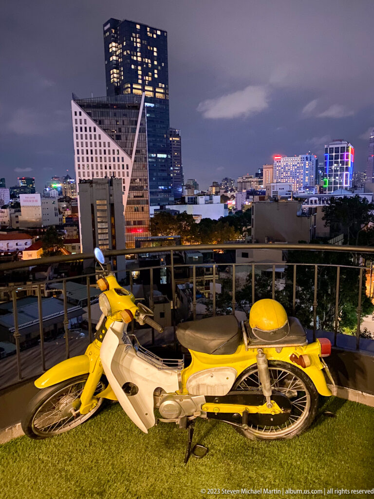 Yellow vintage step-through motorcycle on rooftop turf with the city skyline at night.