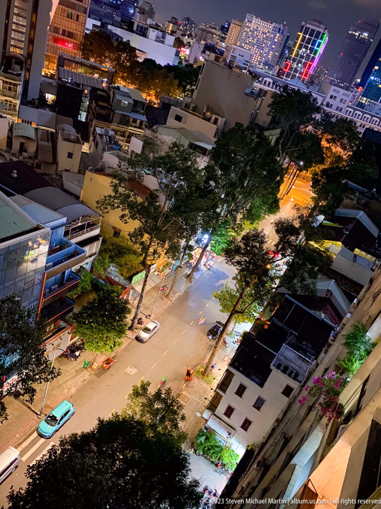 High-angle nighttime view of a lit street, trees, traffic, and city buildings.