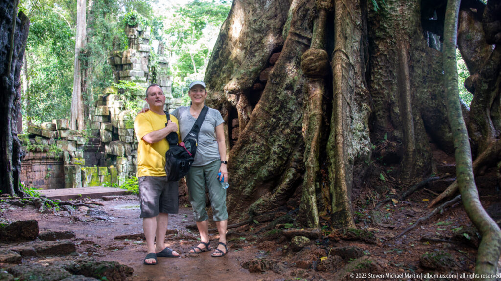 Two visitors standing between enormous tree buttress roots at Angkor Thom.