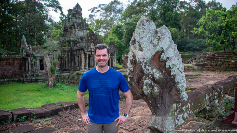 Man in blue posing beside a weathered stone guardian at Angkor Thom.
