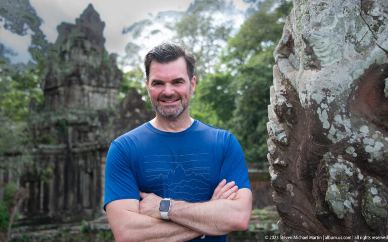 Closer portrait of man in blue beside the guardian sculpture at Angkor Thom.