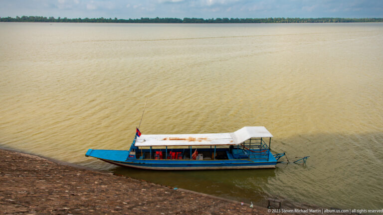 Long covered passenger boat crossing brown reservoir water at Angkor Thom.