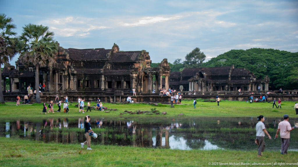 Daylight frontal view of the western façade with visitors crossing grass beside the pool at Angkor Wat.
