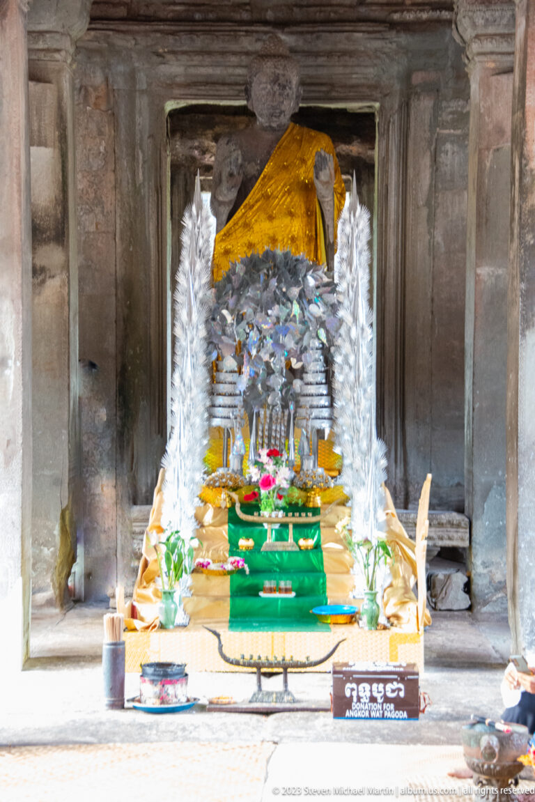 Active shrine with a dark multi-armed sacred image, flowers, and bright offerings at Angkor Wat.
