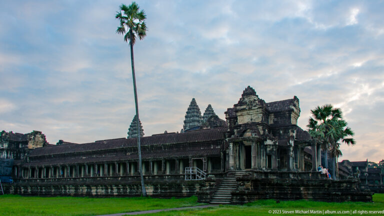 Wide side view of the outer gallery and tower with a tall palm rising beside the structure at Angkor Wat.