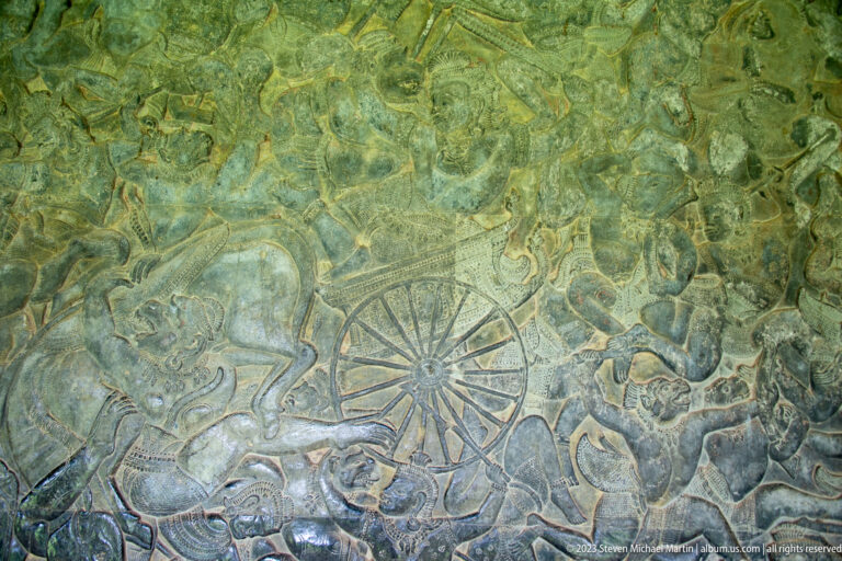 Large wheel-like circular motif amid figures in a green-stained relief at Angkor Wat.