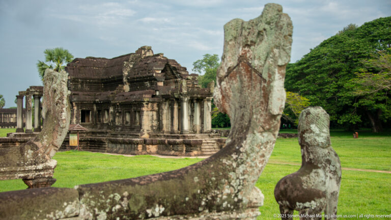 Weathered naga balustrade head in foreground with long gallery across the lawn at Angkor Wat.