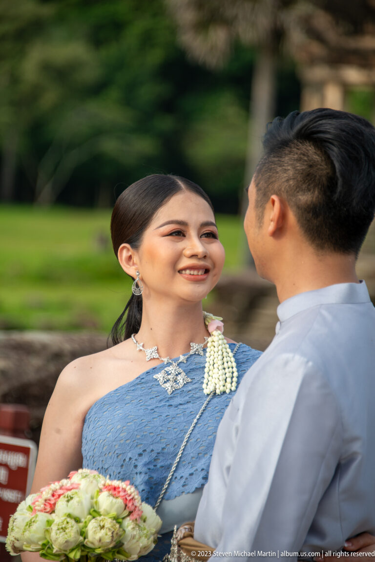 Young woman in formal blue traditional dress smiling toward a man at Angkor Wat.