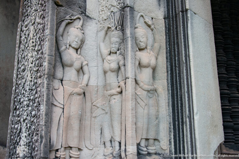 Three standing devatas carved in pale sandstone beside a decorated vertical border at Angkor Wat.