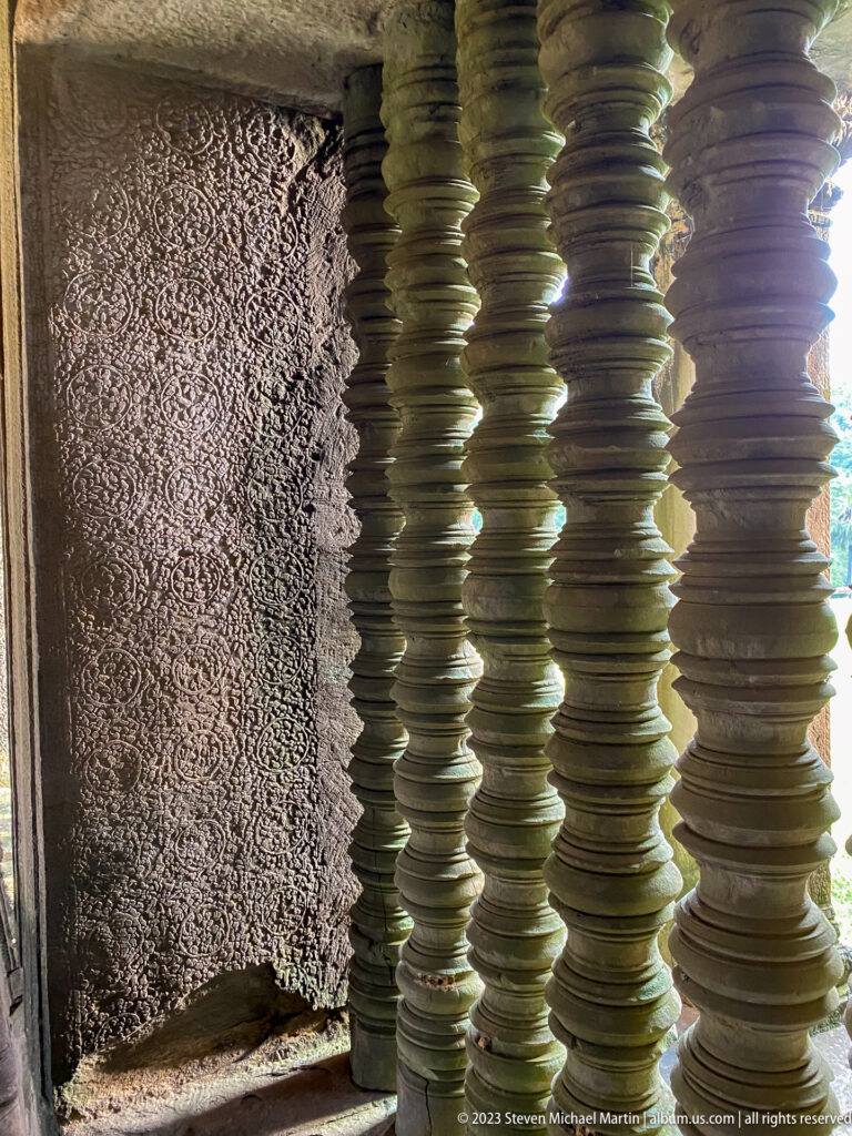 Turned stone window balusters lit from the side beside a rough wall at Angkor Wat.