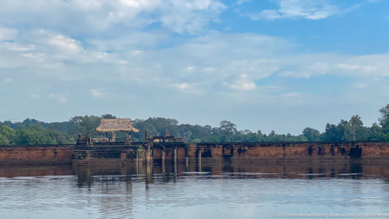 Distant waterside pavilion and tree line reflected across broad calm water at Angkor Wat.