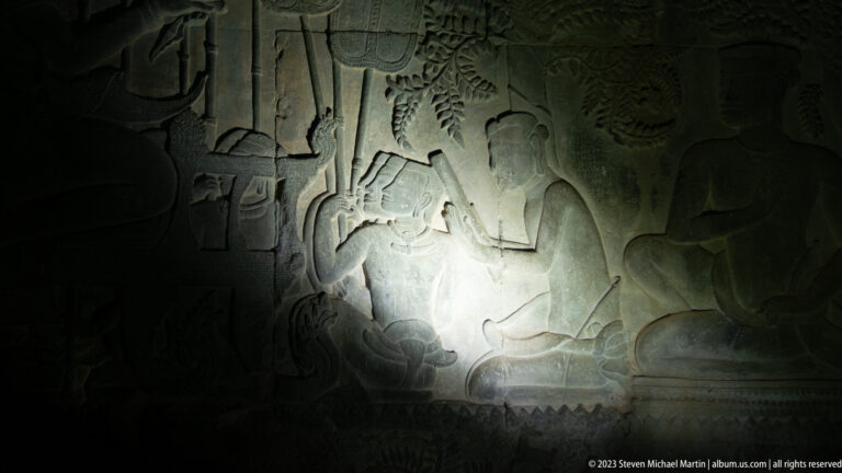 Dark bas-relief illuminated by a circular patch of light showing a figure amid surrounding forms at Angkor Wat.