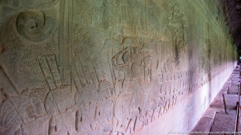 Long gallery relief with ranks of small standing figures and a bright diagonal patch of light at Angkor Wat.