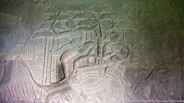 Bas-relief with a long boat or chariot-like form beneath standing figures and vertical standards at Angkor Wat.