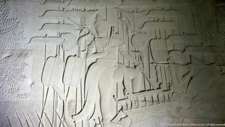 Processional relief with tall figures and a long horizontal vehicle or boat form at Angkor Wat.